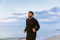 Homem barbudo confiante em sportswear correndo na areia no mar ao pôr do sol — Fotografia de Stock