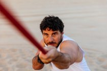 Fiducioso uomo barbuto in abbigliamento sportivo tirando corda mentre si esercita sulla spiaggia di sabbia — Foto stock