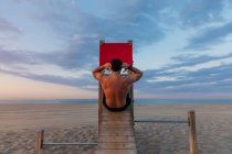 Back view of shirtless muscular guy doing abdominal crunches on wooden slide on beach at sunset — Stock Photo