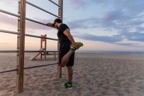 Мускулистый парень в спортивной одежде делает разминку для ног, стоя возле лестницы во время заката на пляже — стоковое фото