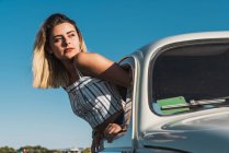 Felice giovane donna in occhiali da sole alla moda guardando fuori dalla finestra dell'auto godendo la luce del sole estiva contro alberi verdi e cielo blu — Foto stock