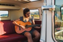 Женщина играет на акустической гитаре, сидя в фургоне ретро во время поездки — стоковое фото