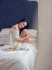 Mujer joven desayunando sentada en la cama - foto de stock