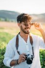 Beau jeune homme en lunettes de soleil souriant joyeusement et tenant appareil photo rétro tout en se tenant debout sur fond flou de campagne étonnante — Photo de stock