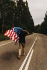 Vista trasera del hombre descalzo sosteniendo la bandera de EE.UU. y corriendo a lo largo de estrecha carretera rural - foto de stock