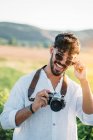 Красивый молодой парень в солнечных очках весело улыбается и держит ретро фотокамеру, стоя на размытом фоне удивительной сельской местности — стоковое фото