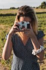 Mulher de vestido tirando foto com câmera retro em pé no fundo da paisagem verde de verão à luz do sol — Fotografia de Stock