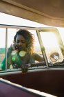 Lovely African-American woman smiling and looking at camera through window of vintage car while spending time in nature on sunny day — Stock Photo