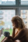 Douce femme nue assise sur le rebord de la fenêtre avec une tasse de café — Photo de stock