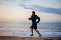 Homme en vêtements de sport courir sur le sable en mer pendant l'entraînement en plein air sur la plage au coucher du soleil — Photo de stock