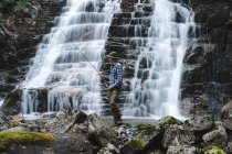 Fischer mit Fischerhut, blauem Hemd und grauer Marschhose, der eine Spinnrute hält, die auf Felsen hinter dem Hang eines Wasserfalls steht — Stockfoto