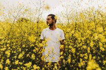 Hombre guapo en camiseta blanca mirando hacia otro lado mientras está de pie entre hermosas flores amarillas - foto de stock