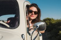 Mujer joven feliz en gafas de sol elegantes mirando por la ventana del coche disfrutando de la luz del sol de verano contra árboles verdes y el cielo azul - foto de stock