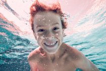 Rothaariges Kind, das im Wasser taucht und vor dem Hintergrund transparenten Wassers in die Kamera blickt — Stockfoto