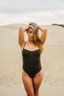 Portrait of young sensual in black swimwear standing on sand with hands in hair — Stock Photo