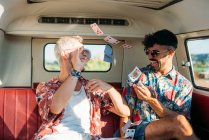 Two cheerful young guys laughing and throwing playing cards while sitting on passenger seat of retro van during trip in nature — Stock Photo