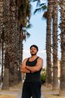 Bearded man in sportswear with arms crossed and looking at camera while standing among palms during outdoor training — Stock Photo