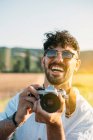 Bello giovane ragazzo in occhiali da sole allegro sorridente e tenendo macchina fotografica retrò mentre in piedi su sfondo sfocato di campagna incredibile — Foto stock