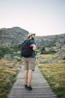 Young man in hat with backpack walking on wooden path in mountains — Stock Photo