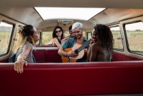 Група молодих людей сміється і слухає красивого хлопця, який грає на акустичній гітарі всередині старовинного фургона в природі — стокове фото
