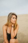 Jovem mulher pensativa em roupa de banho preta sentada na areia e olhando para longe — Fotografia de Stock