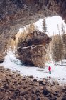 Person mit roter Weste und blauer Hose steht im kanadischen Winterwald mit riesigen braunen Felsen und hohen Tannen am Tag — Stockfoto
