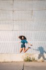 Funky giovane donna afroamericana con i capelli scuri in denim vestiti e scarpe da ginnastica saltando alto sulla strada contro il muro — Foto stock
