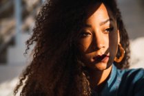 Jeune femme afro-américaine réfléchie avec des cheveux bouclés et un maquillage vif regardant loin avec ombre sur le visage — Photo de stock