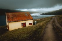 Pequeña casa gruñona con techo oxidado en la orilla del lago en las Islas Feroe en día nublado - foto de stock