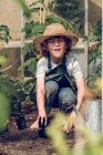 Маленький выразительный мальчик в грязных джинсах и соломенной шляпе саженцы растений в почве в оранжерее — стоковое фото