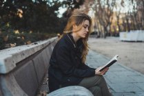 Молодая женщина, сидящая на скамейке в парке и читающая книгу — стоковое фото