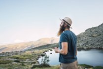 Молодий чоловік у капелюх стоїть біля озера в горах з чашкою і, дивлячись на вид — стокове фото