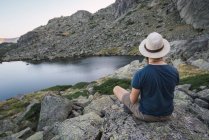 Young man in hat sitting on rocks near lake and looking at view — Stock Photo