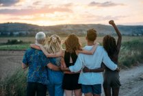 Back view of young men and women embracing each other and admiring beautiful sunset while spending time in nature together — Stock Photo