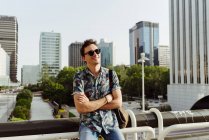 Cheerful young man in sunglasses leaning on handrail and looking away in the city — Stock Photo