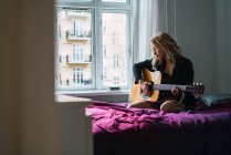 Bionda giovane donna che suona la chitarra sul letto a casa — Foto stock