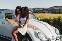 Mulheres multiétnicas na moda em pé perto de carro vintage e ler mapa enquanto viajam juntas no verão — Fotografia de Stock