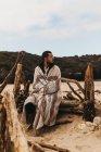 Красивый мужчина, завернутый в декоративное одеяло и смотрящий в сторону, сидя на загвоздках на песчаном побережье — стоковое фото