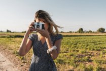 Mulher de vestido tirando foto com câmera retro em pé no fundo da paisagem verde de verão à luz do sol — Fotografia de Stock
