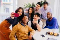Équipe multiraciale debout et assis tout en prenant selfie à la table au bureau. — Photo de stock