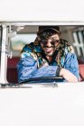 Привлекательный молодой человек в стильном наряде, опирающийся на руль и смотрящий в камеру, сидя в винтажном фургоне во время поездки на природе — стоковое фото