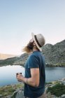 Joven hombre relajado en sombrero de pie cerca del lago con taza y disfrutando de la vista - foto de stock