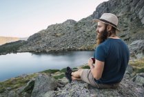 Joven hombre relajado en sombrero sentado en las rocas cerca del lago en las montañas - foto de stock
