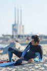 Чоловік у гідрокостюмі сидить з дошкою для серфінгу на пляжі, дивлячись на океан — стокове фото