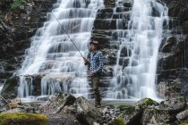 Fischer mit Fischerhut, blauem Hemd und grauer Marschhose, der eine Spinnrute hält, die auf Felsen hinter dem Hang eines Wasserfalls steht — Stockfoto