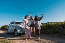 Стильные мультиэтнические женщины делают селфи со смартфоном на сельской дороге с винтажным автомобилем на фоне голубого неба — стоковое фото