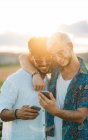 Dos hombres sonrientes abrazando y navegando teléfonos inteligentes mientras están de pie en un hermoso campo juntos - foto de stock