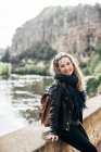 Lovely young female smiling and looking at camera while leaning on concrete border of bridge on blurred background of water and cliff — Stock Photo
