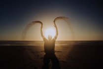 Silhouette d'une personne méconnaissable jetant du sable sur la plage au coucher du soleil retour éclairé. — Photo de stock