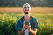 Красивый молодой парень в стильной одежде смеется и просматривает смартфон, стоя на фоне красивой природы в солнечный день — стоковое фото